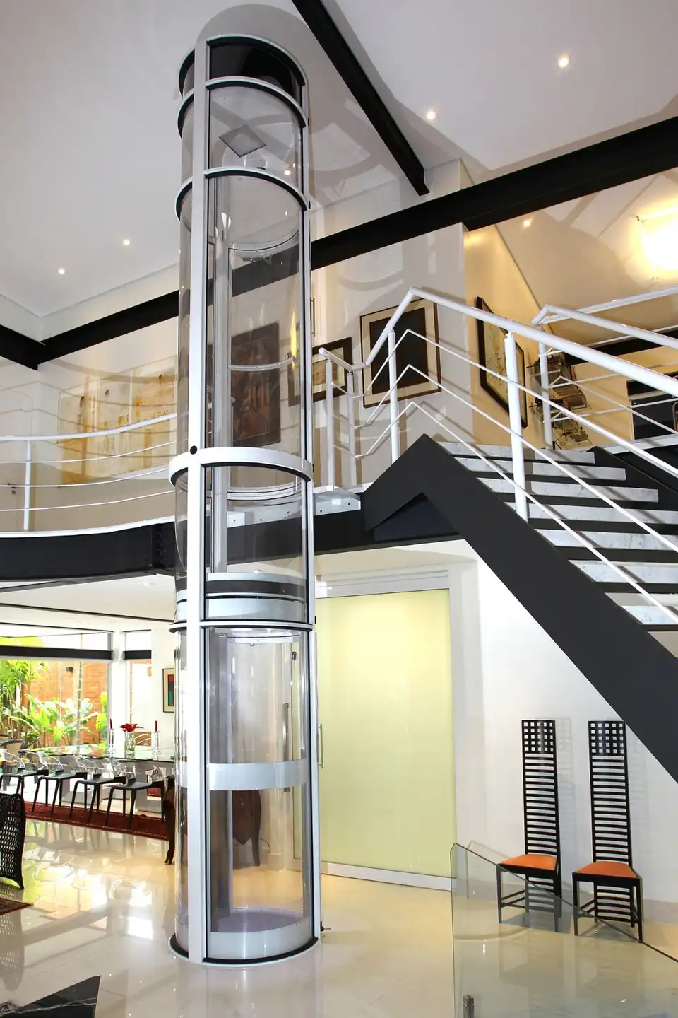 Elevador residencial transparente conectando diferentes pavimentos junto a uma escada arquitetônica