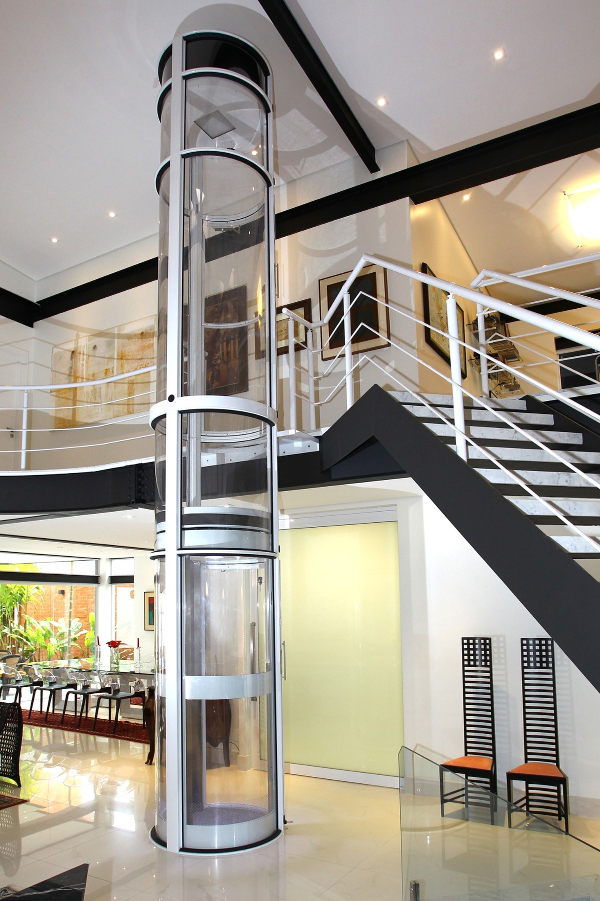 Elevador residencial transparente conectando diferentes pavimentos junto a uma escada arquitetônica