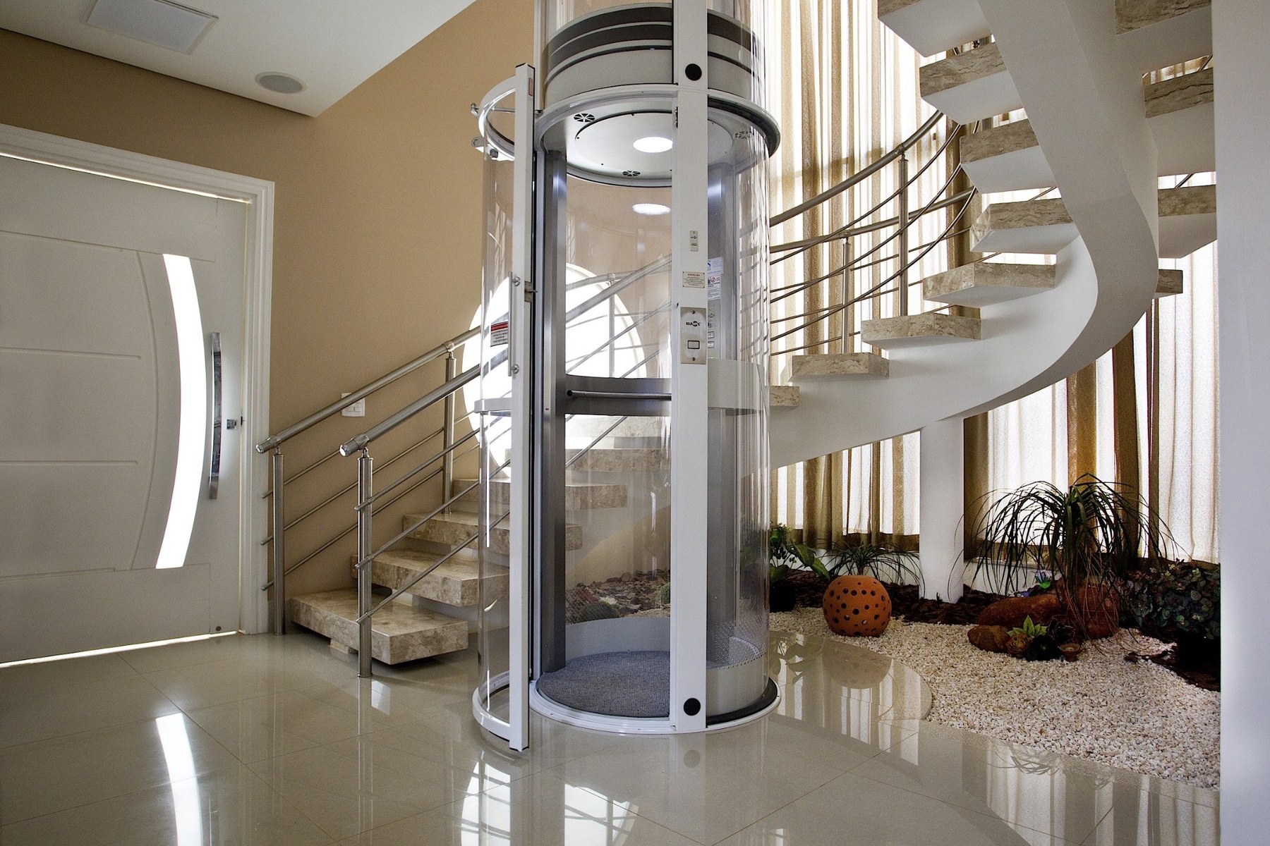 Elevador residencial transparente ao lado de uma escada curva em uma residência